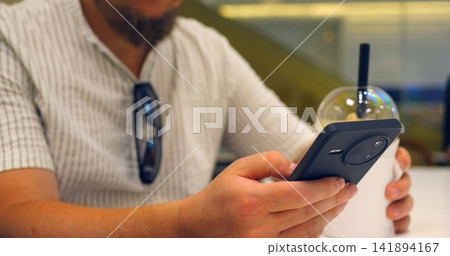 Man holding smartphone and takeaway drink in cafe, using mobile payment at counter with close up hands and cup, wearing casual striped shirt, modern contactless purchase in progress Man holding smartphone and takeaway drink in cafe, using mobile payment at counter with close up hands and cup, wearing casual striped shirt, modern contactless purchase in progress 141894167