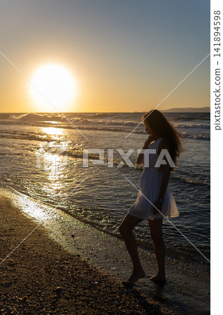 Woman in white dress standing on sandy beach at golden sunset Woman in white dress standing on sandy beach at golden sunset 141894598