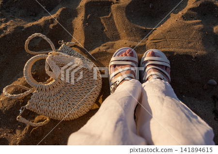 High angle view of female feet in white sandals and woven bag on sand High angle view of female feet in white sandals and woven bag on sand 141894615