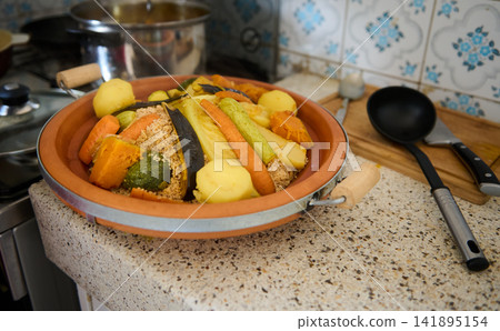 Authentic Moroccan couscous with steamed vegetables served in a clay dish on a kitchen counter 141895154