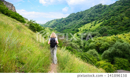 Blonde woman hiking on a mountain trail with a backpack. Female hiker walking along a grassy slope overlooking a lush green valley. Outdoor adventure and active lifestyle concept 141896138