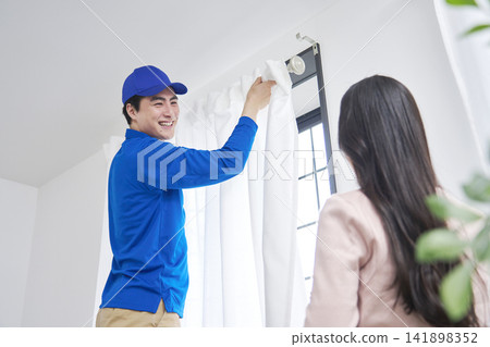The moving company installs the curtains while a woman gives instructions. 141898352