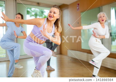 Group of energetic young mature female dancers dancing with knee lift during training together in dance hall Group of energetic young mature female dancers dancing with knee lift during training together in dance hall 141898564