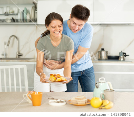 Guy gently hugs girlfriend around waist during preparations for afternoon snack Guy gently hugs girlfriend around waist during preparations for afternoon snack 141898941