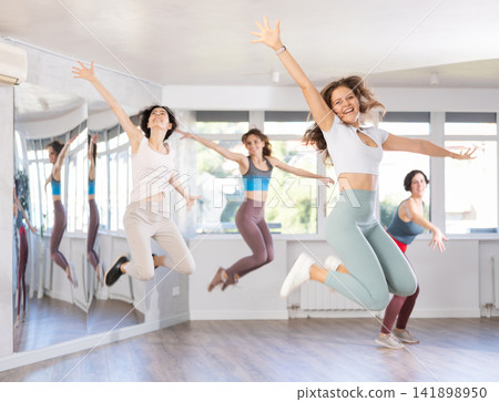 Girls engaged shake leg in dance fitness hall with friends, group of youthful people jumped into air Girls engaged shake leg in dance fitness hall with friends, group of youthful people jumped into air 141898950