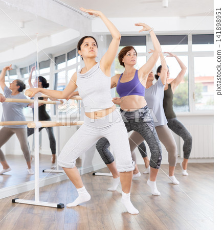 Women doing ballet at group amateurs training session in the studio performs demi plie near the ballet barre, standing in ballet stance Women doing ballet at group amateurs training session in the studio performs demi plie near the ballet barre, standing in ballet stance 141898951
