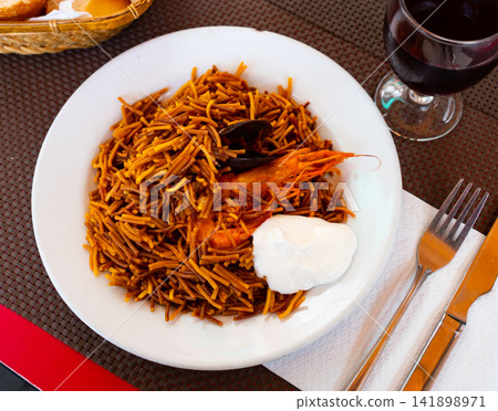 Appetizing traditional Spanish dish made from seafood and noodles served on a plate with aioli sauce on the table in a restaurant Appetizing traditional Spanish dish made from seafood and noodles served on a plate with aioli sauce on the table in a restaurant 141898971