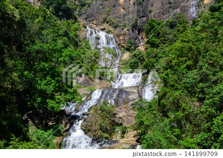 Waterfall cascades down rugged rocky cliffs surrounded by lush green forest in the highlands of Nuwara Eliya, Sri Lanka Waterfall cascades down rugged rocky cliffs surrounded by lush green forest in the highlands of Nuwara Eliya, Sri Lanka 141899709
