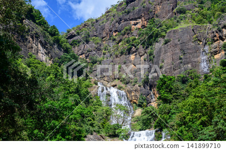 Waterfall cascades down rugged rocky cliffs surrounded by lush green forest in the highlands of Nuwara Eliya, Sri Lanka Waterfall cascades down rugged rocky cliffs surrounded by lush green forest in the highlands of Nuwara Eliya, Sri Lanka 141899710