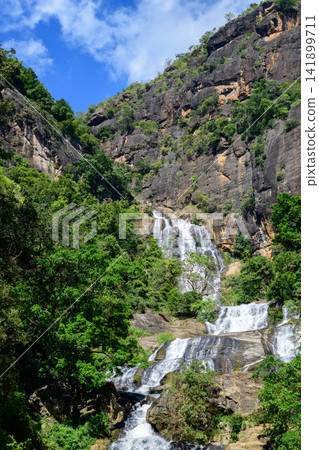Waterfall cascades down rugged rocky cliffs surrounded by lush green forest in the highlands of Nuwara Eliya, Sri Lanka Waterfall cascades down rugged rocky cliffs surrounded by lush green forest in the highlands of Nuwara Eliya, Sri Lanka 141899711