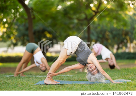 Diligent women practicing feet apart forward bend pose of yoga on mat at city park together Diligent women practicing feet apart forward bend pose of yoga on mat at city park together 141902472