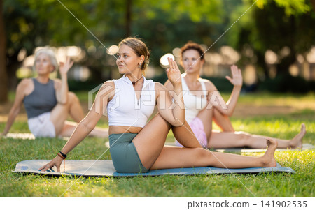 Diligent young woman practicing lord of the fishes pose of yoga on mat in light green park at daytime Diligent young woman practicing lord of the fishes pose of yoga on mat in light green park at daytime 141902535