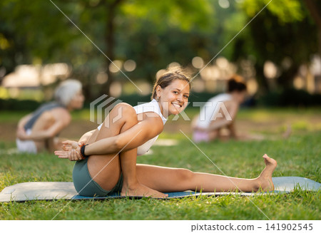 Smiling girl performing seated asana Marichyasana during yoga class in park Smiling girl performing seated asana Marichyasana during yoga class in park 141902545