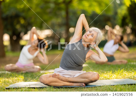 Happy old lady and class of calm sports women in activewear stretching muscles and doing tilts while sitting in lotus pose on mats during yoga training together at city park Happy old lady and class of calm sports women in activewear stretching muscles and doing tilts while sitting in lotus pose on mats during yoga training together at city park 141902556