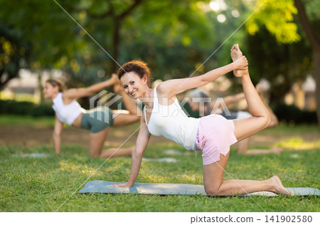 Woman with group performing Kneeling Bow yoga pose in park Woman with group performing Kneeling Bow yoga pose in park 141902580