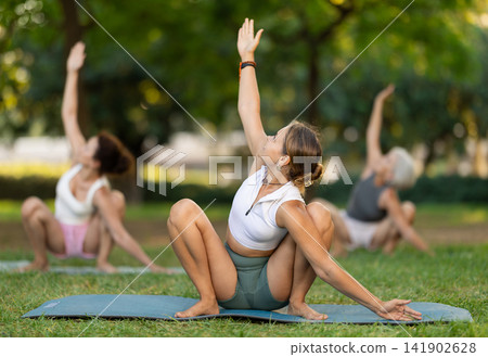 Active young girl teacher performs garland pose with group in green park Active young girl teacher performs garland pose with group in green park 141902628