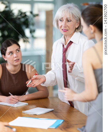 Focused mature female office worker standing with pen in hands near table where sitting colleagues with papers and emotional discussing problems in office Focused mature female office worker standing with pen in hands near table where sitting colleagues with papers and emotional discussing problems in office 141902700