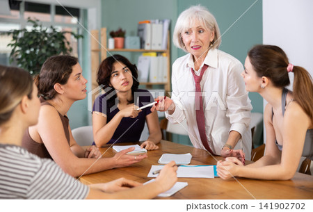 Focused mature female office worker standing with pen in hands near table where sitting colleagues with papers and emotional discussing problems in office Focused mature female office worker standing with pen in hands near table where sitting colleagues with papers and emotional discussing problems in office 141902702