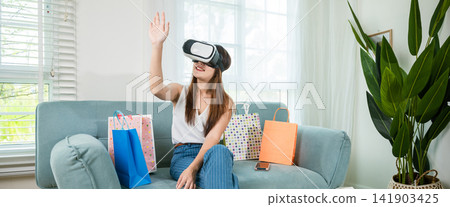 Happy woman customer with shopping bags around wear VR headset on sofa touching air Happy woman customer with shopping bags around wear VR headset on sofa touching air 141903425