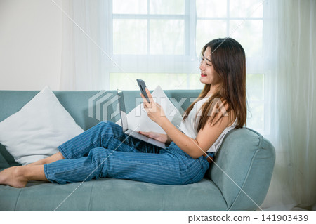 woman using smart mobile phone while sitting on sofa in living room at home with laptop computer woman using smart mobile phone while sitting on sofa in living room at home with laptop computer 141903439