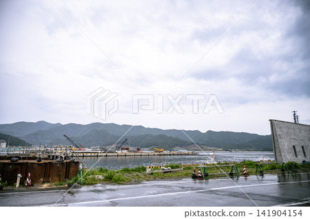 在陰雲密佈的天空下,平靜的海灣和港口建設工程透過為應對海嘯而新建的海堤隱約可見。 在陰雲密佈的天空下,平靜的海灣和港口建設工程透過為應對海嘯而新建的海堤隱約可見。 141904154