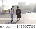 Male students walking on campus rooftop, graduate wearing gown and cap holding diploma and phone 141907784