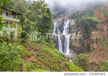 Ramboda Waterfall near Nuwara Eliya town in Sri Lanka. Majestic multi-tiered waterfall cascades through lush green mountainside with a charming hillside dwelling nearby Ramboda Waterfall near Nuwara Eliya town in Sri Lanka. Majestic multi-tiered waterfall cascades through lush green mountainside with a charming hillside dwelling nearby 141907925