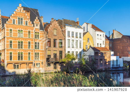 Ghent town in Belgium, Europe. Historic European Canal Houses Reflecting in Water with Reed Beds Under a Clear Blue Sky Ghent town in Belgium, Europe. Historic European Canal Houses Reflecting in Water with Reed Beds Under a Clear Blue Sky 141907928
