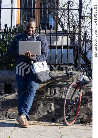 African American man leaning on stone wall by sidewalk using laptop with messenger bag, road bike African American man leaning on stone wall by sidewalk using laptop with messenger bag, road bike 141908101