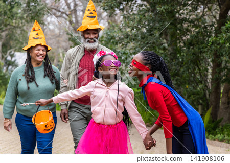 African American family walking along paved garden path wearing pumpkin hats, holding candy bucket 141908106
