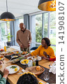Family gathering around long wooden dining table at home, host carving roasted chicken 141908107