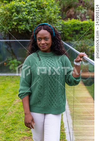 African American woman posing at glass rail beside path in garden wearing green knit teal headband African American woman posing at glass rail beside path in garden wearing green knit teal headband 141908114