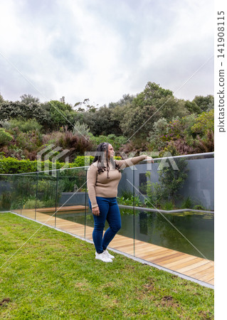 Woman standing on grassy lawn beside glass railing and wooden deck near reflective pond, copy space Woman standing on grassy lawn beside glass railing and wooden deck near reflective pond, copy space 141908115