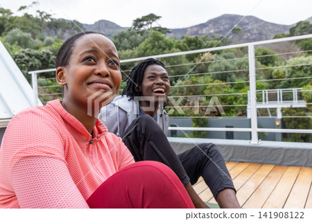 African American couple sitting on rooftop deck by cable railing on yoga mat in sportswear smiling African American couple sitting on rooftop deck by cable railing on yoga mat in sportswear smiling 141908122