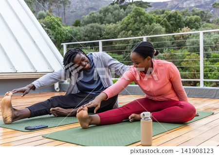 African American couple stretching on rooftop terrace in activewear with mats, phone, water bottle 141908123