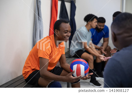 Diverse male volleyball teammates sitting on bench in locker room holding tricolor volleyball Diverse male volleyball teammates sitting on bench in locker room holding tricolor volleyball 141908127