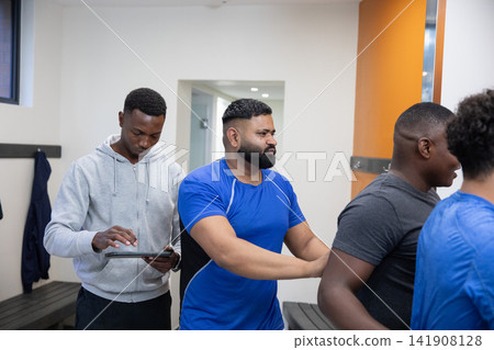 Diverse male teammates helping in sportswear, placing hands on back in locker room with tablet 141908128
