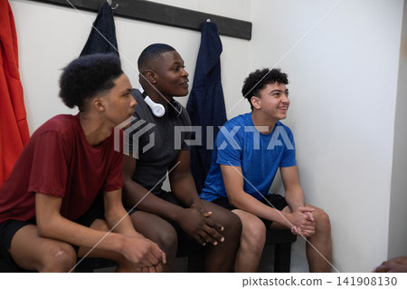 Diverse male teens sitting on wooden bench in locker room wearing shirts and headset near jackets Diverse male teens sitting on wooden bench in locker room wearing shirts and headset near jackets 141908130