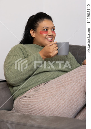 Woman in twenties sitting on gray sofa wearing green sweater and red under-eye patches holding mug 141908140