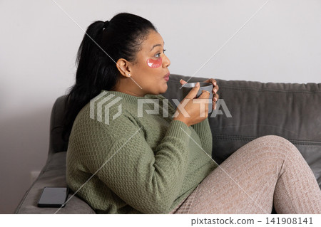 African American woman sitting on grey sofa at home cradling mug wearing olive sweater eye-patches African American woman sitting on grey sofa at home cradling mug wearing olive sweater eye-patches 141908141
