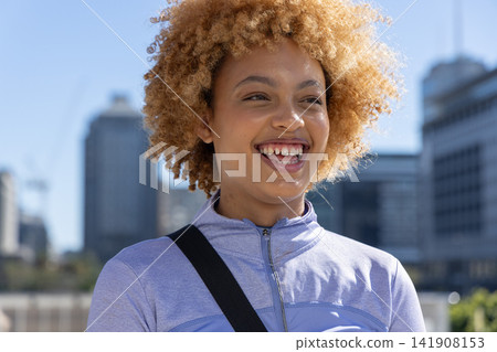 African American female smiling wearing light purple zip-up jacket and black bag strap in city African American female smiling wearing light purple zip-up jacket and black bag strap in city 141908153