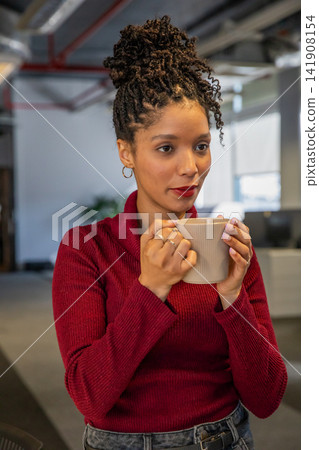 African American woman standing in modern office wearing red knit sweater, jeans, holding beige mug African American woman standing in modern office wearing red knit sweater, jeans, holding beige mug 141908154