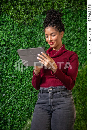 African American woman wearing burgundy turtleneck and jeans, holding gray tablet by hedge wall African American woman wearing burgundy turtleneck and jeans, holding gray tablet by hedge wall 141908156