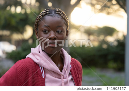 African American woman standing in park wearing pink hoodie and red jacket with braided blonde hair 141908157