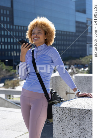 African American woman in activewear holding phone at mouth, crossbody bag and smartwatch on plaza African American woman in activewear holding phone at mouth, crossbody bag and smartwatch on plaza 141908158