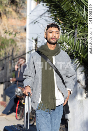 Man walking path by wall wearing beanie and scarf pulling rolling suitcase carrying crossbody bag Man walking path by wall wearing beanie and scarf pulling rolling suitcase carrying crossbody bag 141908159