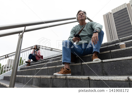 Diverse pedestrians sitting on overpass steps, man with headphones holding smartphone, cup nearby 141908172