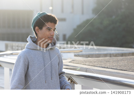 African American teenage male standing on rooftop by handrail, wearing gray hoodie and green beanie African American teenage male standing on rooftop by handrail, wearing gray hoodie and green beanie 141908173