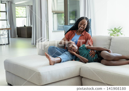 Mother and daughter lying on L-shaped sofa at home, woman in rust shirt caressing youth's head 141908180
