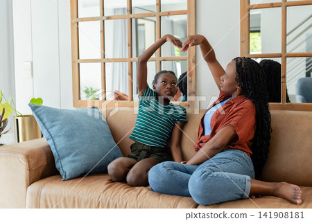 African mother and child sitting on tan sofa making heart overhead in rust top, green-striped tee African mother and child sitting on tan sofa making heart overhead in rust top, green-striped tee 141908181